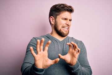 Young handsome blond man with beard and blue eyes wearing casual sweater disgusted expression, displeased and fearful doing disgust face because aversion reaction. With hands raised