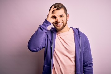 Young blond man with beard and blue eyes wearing purple sweatshirt over pink background doing ok gesture with hand smiling, eye looking through fingers with happy face.