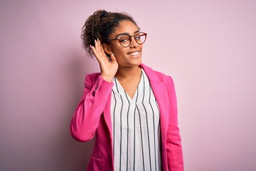 Beautiful african american businesswoman wearing jacket and glasses over pink background smiling with hand over ear listening an hearing to rumor or gossip. Deafness concept.