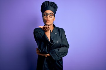 Young african american chef girl wearing cooker uniform and hat over purple background looking at the camera blowing a kiss with hand on air being lovely and sexy. Love expression.