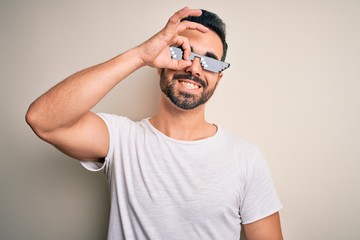 Young handsome man with beard wearing funny thug life sunglasses over white background doing ok gesture with hand smiling, eye looking through fingers with happy face.