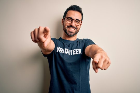 Handsome man with beard wearing t-shirt with volunteer message over white background pointing to you and the camera with fingers, smiling positive and cheerful