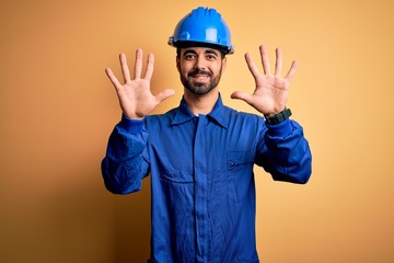 Mechanic man with beard wearing blue uniform and safety helmet over yellow background showing and...