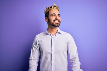 Obraz premium Young handsome man with beard wearing golden crown of king over purple background looking away to side with smile on face, natural expression. Laughing confident.