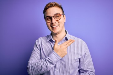 Young handsome redhead man wearing casual shirt and glasses over purple background cheerful with a smile of face pointing with hand and finger up to the side with happy and natural expression on face