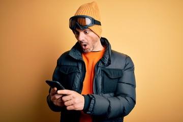 Young handsome skier man wearing ski goggles using smartphone over yellow background scared in...