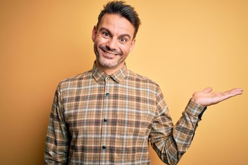 Young handsome man wearing casual shirt standing over isolated yellow background smiling cheerful presenting and pointing with palm of hand looking at the camera.