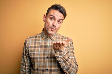Young handsome man wearing casual shirt standing over isolated yellow background looking at the camera blowing a kiss with hand on air being lovely and sexy. Love expression.