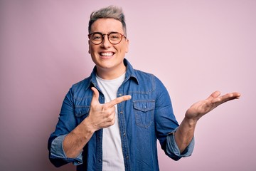 Young handsome modern man wearing glasses and denim jacket over pink isolated background amazed and smiling to the camera while presenting with hand and pointing with finger.