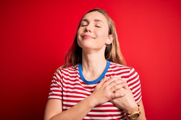 Young beautiful blonde woman wearing casual striped t-shirt over isolated red background smiling with hands on chest with closed eyes and grateful gesture on face. Health concept.