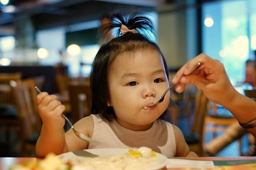 A cute Asian girl eating salad at a restaurant with her mom feeding her using fork and spoon.