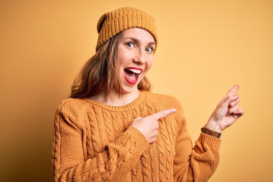 Young beautiful blonde woman wearing casual sweater and wool cap over yellow background smiling and looking at the camera pointing with two hands and fingers to the side.