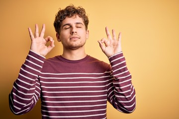 Fototapeta premium Young blond handsome man with curly hair wearing casual striped sweater relax and smiling with eyes closed doing meditation gesture with fingers. Yoga concept.