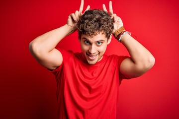 Fototapeta na wymiar Young blond handsome man with curly hair wearing casual t-shirt over red background Posing funny and crazy with fingers on head as bunny ears, smiling cheerful