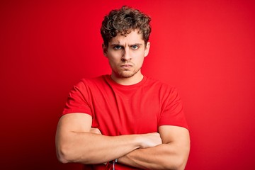 Young blond handsome man with curly hair wearing casual t-shirt over red background skeptic and...