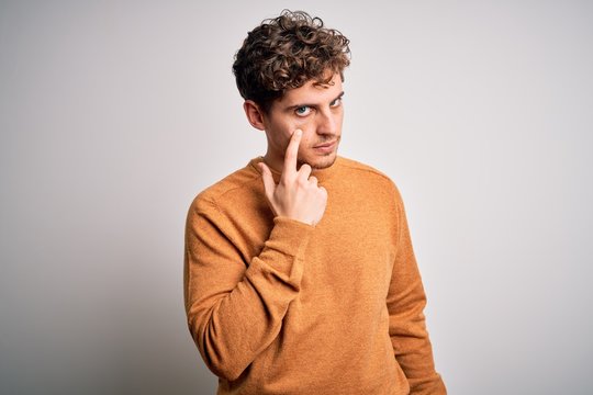 Young blond handsome man with curly hair wearing casual sweater over white background Pointing to the eye watching you gesture, suspicious expression