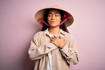 Fototapeta premium Young beautiful woman wearing traditional conical asian hat over isolated pink background smiling with hands on chest with closed eyes and grateful gesture on face. Health concept.