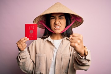 Young thai woman wearing traditional conical asian hat holding japan japanese passport annoyed and frustrated shouting with anger, crazy and yelling with raised hand, anger concept