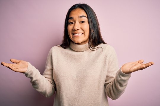 Young beautiful asian woman wearing casual turtleneck sweater over pink background clueless and confused expression with arms and hands raised. Doubt concept.