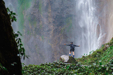 Happy satisfied long haired woman wearing black cap standing on the cliff to watch Tumpak Sewu waterfall from above and enjoy scenic green nature beauty.