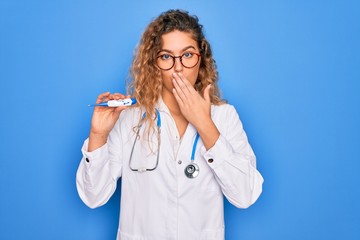 Young beautiful blonde doctor woman with blue eyes wearing stethoscope holding thermometer cover mouth with hand shocked with shame for mistake, expression of fear, scared in silence, secret concept