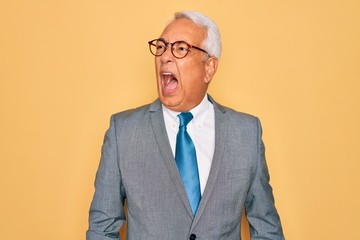 Middle age senior grey-haired handsome business man wearing glasses over yellow background angry and mad screaming frustrated and furious, shouting with anger. Rage and aggressive concept.