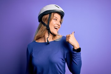 Young beautiful blonde cyclist woman wearing bike security helmet over purple background smiling with happy face looking and pointing to the side with thumb up.