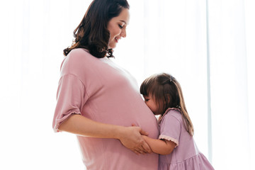 Side view of cute mixed race toddler kissing tummy of happy pregnant woman at home