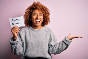Young African American afro woman with curly hair holding paper with fake news message very happy...