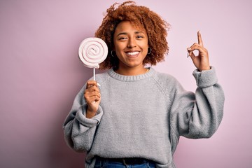 Young beautiful African American afro woman with curly hair eating sweet candy surprised with an idea or question pointing finger with happy face, number one