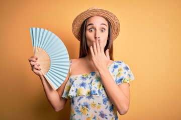 Young tourist woman on vacation wearing summer hat holding hand fan over yellow background cover...