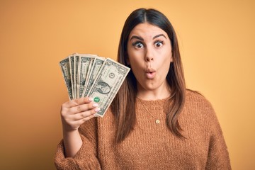 Young beautiful woman holding dollars standing over isolated orange background scared in shock with a surprise face, afraid and excited with fear expression