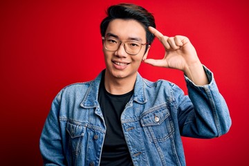 Young handsome chinese man wearing denim jacket and glasses over red background smiling and confident gesturing with hand doing small size sign with fingers looking and the camera. Measure concept.