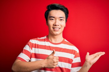 Young handsome chinese man wearing casual striped t-shirt standing over red background Showing palm hand and doing ok gesture with thumbs up, smiling happy and cheerful