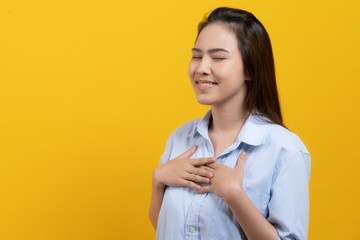 Appreciated expression. Pleased asian woman keeping hands on chest and closing her eyes isolated on yellow background studio shot.