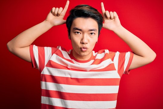 Young handsome chinese man wearing casual striped t-shirt standing over red background doing funny gesture with finger over head as bull horns