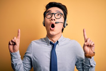Young handsome chinese call center agent man wering glasses using headset amazed and surprised...