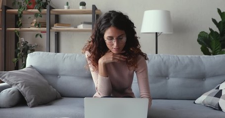 Millennial woman using laptop computer working online sitting on sofa at home office. Smart young lady doing online job thinking of internet research, browsing web, e learning with pc and papers.