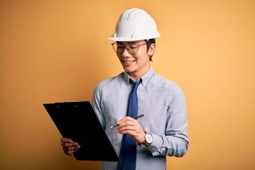 Young handsome chinese engineer man wearing safety helmet holding clipboard with a happy face standing and smiling with a confident smile showing teeth