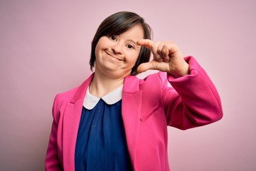 Young down syndrome business woman over pink background smiling and confident gesturing with hand doing small size sign with fingers looking and the camera. Measure concept.