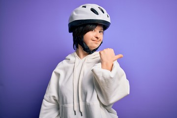 Young down syndrome cyclist woman wearing security bike helmet over purple background smiling with happy face looking and pointing to the side with thumb up.