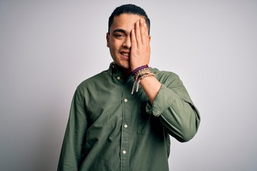 Young brazilian man wearing casual shirt standing over isolated white background covering one eye with hand, confident smile on face and surprise emotion.