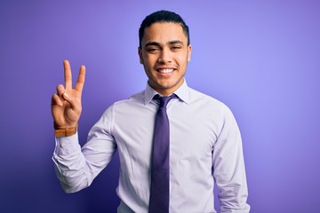 Young brazilian businessman wearing elegant tie standing over isolated purple background showing and pointing up with fingers number two while smiling confident and happy.