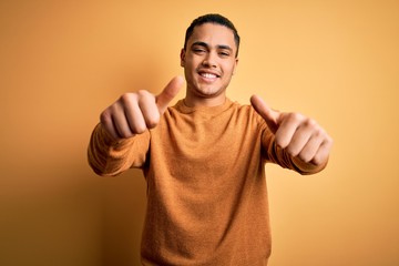 Young brazilian man wearing casual sweater standing over isolated yellow background approving doing positive gesture with hand, thumbs up smiling and happy for success. Winner gesture.