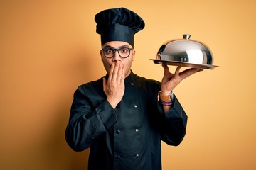 Young brazilian chef man wearing cooker uniform and hat holding tray with dome cover mouth with hand shocked with shame for mistake, expression of fear, scared in silence, secret concept