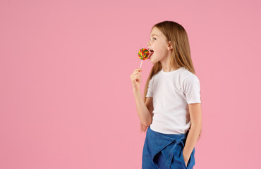 Portrait of a surprised little girl holding heart shaped lollipop and looking at camera isolated over pink background.
