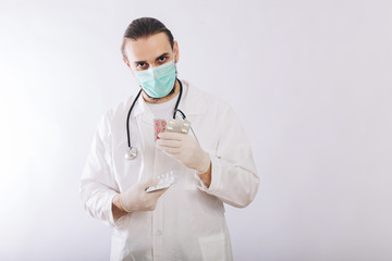 A doctor in a white coat, a medical mask and gloves holds pills. Types of drugs. Medical worker on a white background. Studio photo