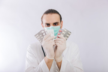 A doctor in a white coat, a medical mask and gloves holds pills. Types of drugs. Medical worker on a white background. Studio photo