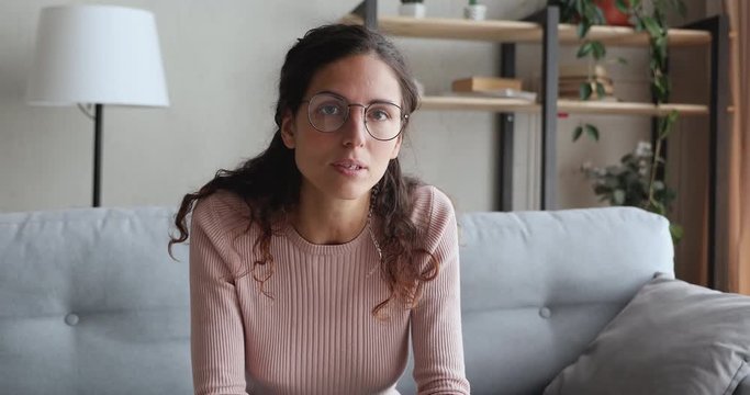 Confident young woman speaking to camera sitting on sofa at home. Female online teacher, blogger, counselor recording podcast for social media channel, making conference video call talking to webcam.