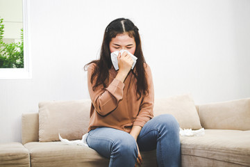 An Asian woman has a headache and a runny nose sitting on the sofa in her house. Concept of personal health care, surveillance of influenza, coronavirus